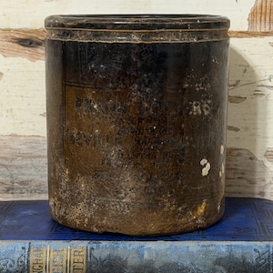 May include: A dark brown cylindrical stoneware crock with a glazed finish and a weathered texture. The front features faded black text that reads Frank Cooper's Seville Marmalade, with additional illegible text below the brand name.