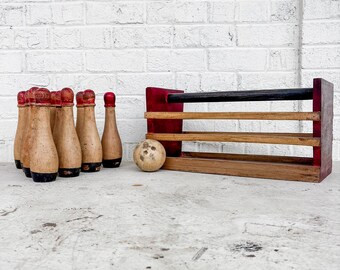 May include: A vintage wooden bowling set with six pins, a ball, and a rack. The pins are tan with red tops and black bases. The ball is light brown with dark spots. The rack is red and brown.