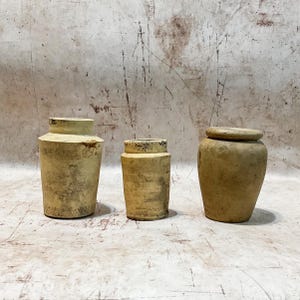 May include: Three antique, cylindrical pottery vessels with a weathered, aged appearance. The vessels are in varying sizes, with a light beige color and dark accents. The vessel on the right has a lid. The background is a neutral, textured surface.
