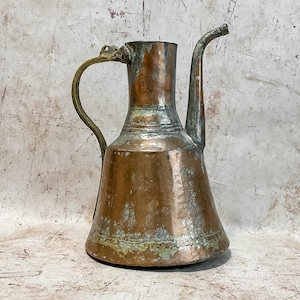 May include: Antique copper pitcher with a curved spout and a brass handle. The pitcher has a wide base that tapers to a narrower neck. The copper has a patina, with areas of green and brown. The handle is attached near the spout and the top of the pitcher.