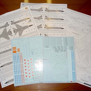 Può includere: Un set di quattro fogli stampati di istruzioni e decalcomanie per un modello di aereo. I fogli sono bianchi con testo e grafica neri e blu. Le decalcomanie sono stampate su plastica trasparente e presentano stelle e numeri rossi, bianchi e blu.