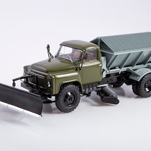 Könnte beinhalten: Ein grüner Spielzeug-LKW mit einem Schneepflug und einem Streueraufsatz. Der LKW hat schwarze Räder und einen schwarzen Schneepflug. Der Streuer ist grau und hat ein Metallgitter.