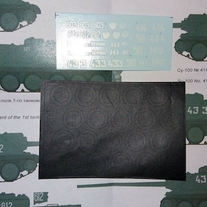 Può includere: Un foglio di decalcomanie per modellismo con numeri e simboli stampati su di essi. Le decalcomanie sono per un modello di veicolo militare. Le decalcomanie sono su uno sfondo bianco.