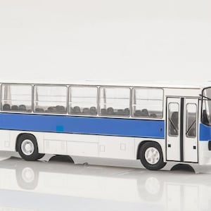 May include: A white and blue scale model bus featuring a rectangular body, large clear windows, two sets of double doors on the side, black windshield wipers, and black tires. The exterior is painted with a horizontal blue stripe along the middle.