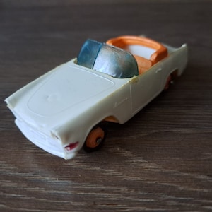 Può includere: Macchina giocattolo bianca con interni arancioni e finestrini colorati di blu. L'auto ha un paraurti cromato e ruote nere.