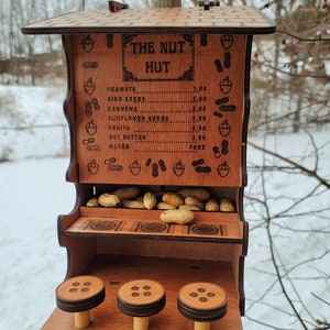 May include: A wooden bird feeder shaped like a hut with a sign that reads "The Nut Hut". The feeder has a price list for different types of bird food, including peanuts, bird seeds, cashews, sunflower seeds, fruits, nut butter, and water. The feeder has a shelf with peanuts inside and three small wooden perches.