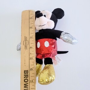May include: A Mickey Mouse plush toy, approximately 7 inches tall, with a black head and ears, a red sequined shorts, and gold sequined feet. The arms have silver glittered hands. The toy has a white face and a black tail.