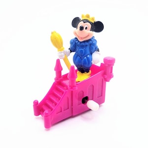 May include: A vintage Mickey Mouse toy figure wearing a blue outfit and a gold crown, holding a yellow broom. The figure stands on a pink castle-like structure with a winding mechanism. The toy is made of plastic.