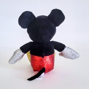 May include: A plush Mickey Mouse toy viewed from the back. The toy has black ears, a black body, and silver glittery gloves. The shorts are red with glitter, and the belly is gold. A black tail extends from the back.