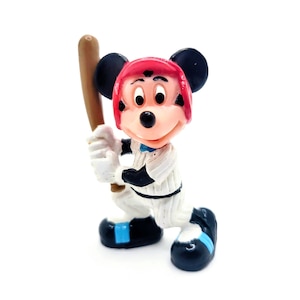 Puede incluir: Una figura de Mickey Mouse con uniforme de b&eacute;isbol, listo para batear con un bate de madera. La figura lleva un casco rojo, un uniforme blanco y azul y zapatos negros. Sobre un fondo blanco.