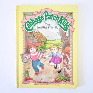May include: A yellow hardcover book titled Cabbage Patch Kids The Just-Right Family, featuring a watercolor illustration of two children running through a forest, with text by Larry Callen and pictures by Judy Gailen, published by Parker Brothers.