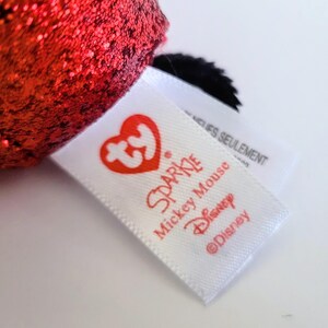 May include: Close-up of a white tag with red text that reads "Sparkle Mickey Mouse Disney &copy;Disney". The tag has a red heart-shaped logo. A red, glittery object is visible in the background.