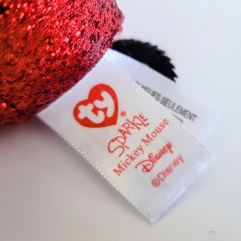 May include: Close-up of a white tag with red text that reads "Sparkle Mickey Mouse Disney &copy;Disney". The tag has a red heart-shaped logo. A red, glittery object is visible in the background.