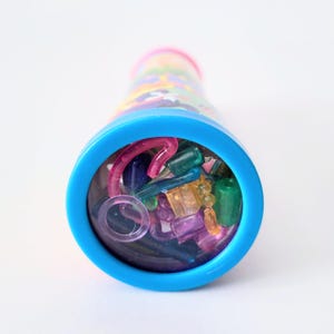 Op de afbeelding: Een kleurrijke caleidoscoop met een blauw oculair en een roze rand. De buis is versierd met een patroon van verschillende kleuren. De caleidoscoop is gevuld met kleine, kleurrijke, doorschijnende vormen in tinten roze, groen, blauw en oranje.