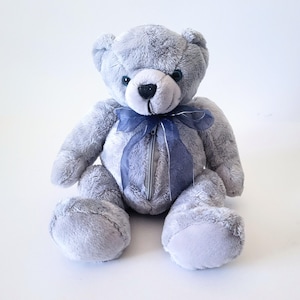 May include: A plush teddy bear covered in soft, light gray fur with dark, round eyes and a black stitched nose. A sheer, dark blue ribbon is tied in a bow around its neck, and a vertical zipper is integrated into the center of its torso.