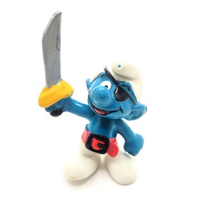 May include: A blue Smurf figurine dressed as a pirate, holding a silver sword with a yellow handle. The Smurf wears a white hat, a black eye patch, a red belt with a black buckle, and white shoes. The figurine is on a white background.