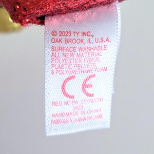 May include: Close-up of a white tag with red text and a red CE symbol. The tag includes the text "2023 TY INC., OAK BROOK, IL, U.S.A." and "HANDMADE IN CHINA". The tag is attached to a red, glittery fabric.