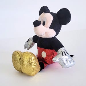 May include: A plush Mickey Mouse toy with black fur, a pink face, and large black ears. The shorts are red with white polka dots, and the gloves are silver. The shoes are gold, and the toy is sitting.