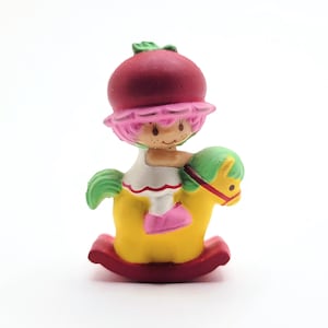 May include: A vintage Strawberry Shortcake figurine riding a yellow rocking horse. The figurine has a red berry hat, pink hair, and a white dress. The rocking horse has green hair and a red base.