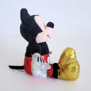 May include: A plush Mickey Mouse toy in a seated position. The toy features black ears, a black body, red sequined shorts, silver sequined gloves, and gold sequined shoes. The face is pink and white with a small smile.