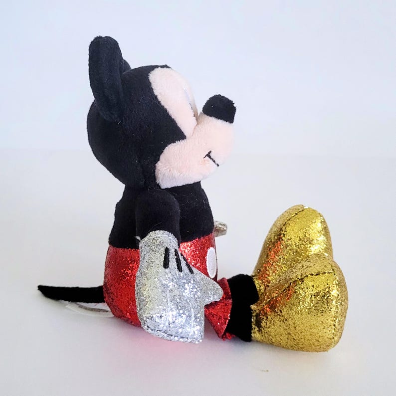 May include: A plush Mickey Mouse toy in a seated position. The toy features black ears, a black body, red sequined shorts, silver sequined gloves, and gold sequined shoes. The face is pink and white with a small smile.