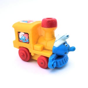 May include: A yellow toy train with red wheels and a red smokestack. The front of the train is a blue Smurf character wearing a white hat. A Smurf character is also visible in a window on the side of the train.