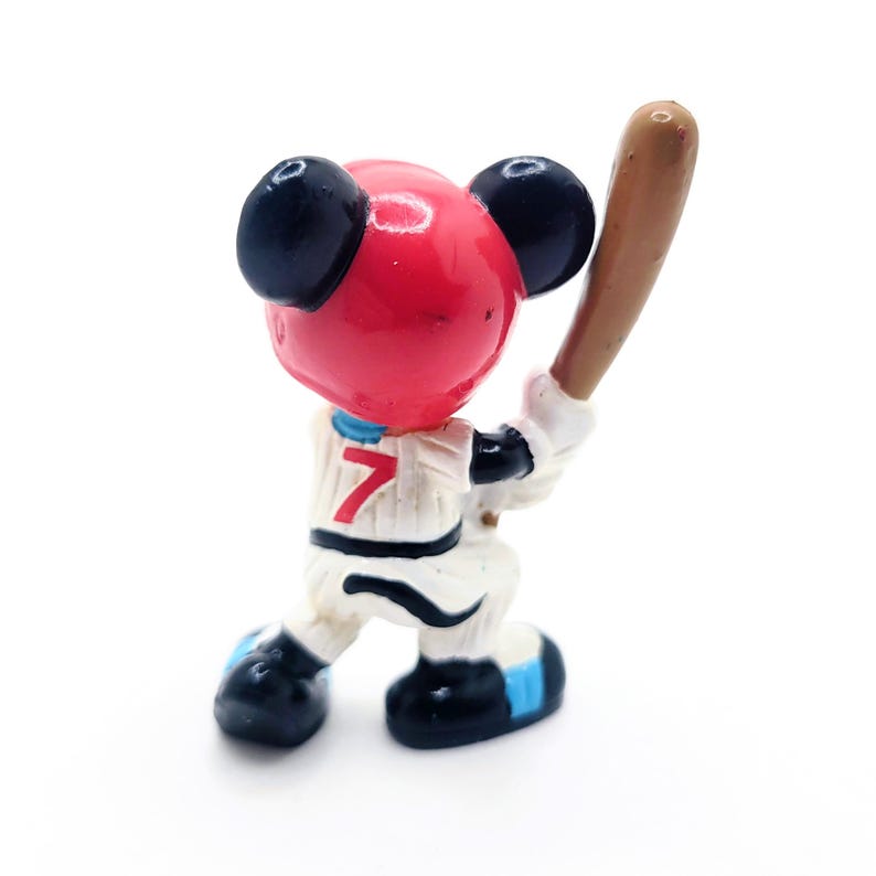 Puede incluir: Peque&ntilde;a figura de un personaje de dibujos animados con uniforme de b&eacute;isbol, sosteniendo un bate. La figura tiene una cabeza roja, orejas negras y un uniforme blanco con el n&uacute;mero 7. El bate es marr&oacute;n y los zapatos son negros con detalles azules.