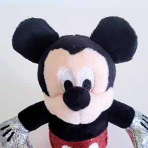 May include: A plush Mickey Mouse toy with black ears, a black nose, and a pink face. The toy has a red sequined bottom and silver glittery hands. The toy is a classic Disney character.