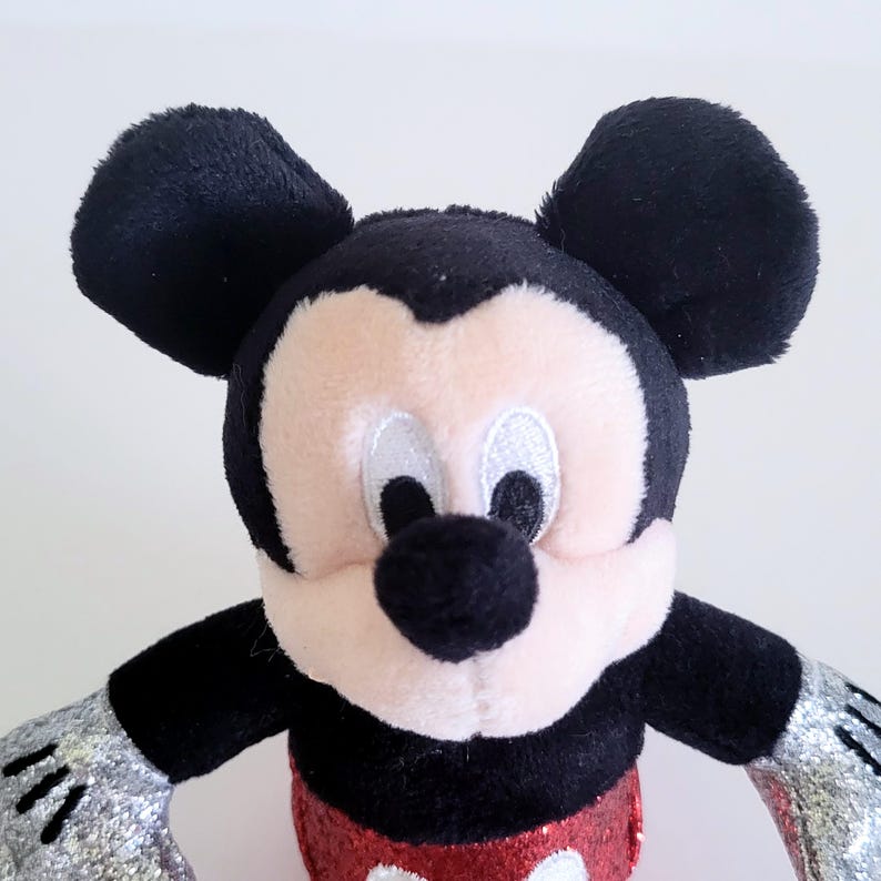 May include: A plush Mickey Mouse toy with black ears, a black nose, and a pink face. The toy has a red sequined bottom and silver glittery hands. The toy is a classic Disney character.