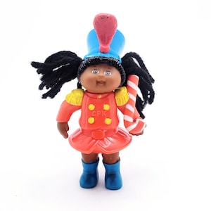 May include: A vintage Cabbage Patch Kids doll in a marching band uniform. The doll has black hair, brown skin, and is wearing a red jacket, skirt, and blue boots. It holds a red and white striped baton and a blue hat with a red plume.
