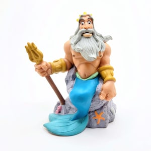 May include: A figurine of a muscular merman with a long white beard, gold crown, and armbands, holding a gold trident. The merman has a blue tail and is perched on a gray rock with an orange starfish.