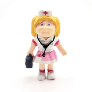 May include: A small figurine of a nurse doll with blonde hair, blue eyes, and a white hat with a red cross. The doll wears a pink and white uniform and holds a black bag and a stethoscope.