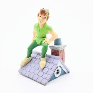May include: A figurine of Peter Pan, dressed in a green tunic and leggings, perched on a roof with a chimney. The roof is light purple with a blue trim. The figurine has a smiling expression.