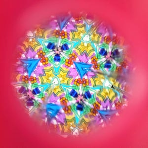 Op de afbeelding: Een caleidoscoopweergave met een levendig, symmetrisch patroon. Het ontwerp bevat blauwe driehoeken, gele stervormen en kleine oranje bloemclusters. De achtergrond is felroze.