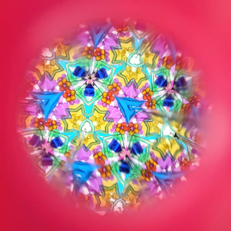 Op de afbeelding: Een caleidoscoopweergave met een levendig, symmetrisch patroon. Het ontwerp bevat blauwe driehoeken, gele stervormen en kleine oranje bloemclusters. De achtergrond is felroze.