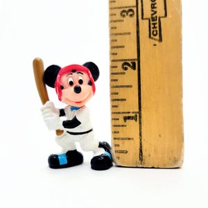 Puede incluir: Una peque&ntilde;a figura de Mickey Mouse con uniforme de b&eacute;isbol, sosteniendo un bate. La figura lleva un casco rojo y un uniforme blanco con detalles negros. Una regla de madera est&aacute; al lado de la figura, con medidas en cent&iacute;metros.