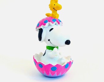 May include: A small figurine of a white beagle with black ears sitting inside a pink eggshell with blue polka dots. A small yellow bird figure is perched on top of the beagle's head. The beagle wears a green collar around its neck.