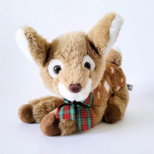 May include: A plush fawn toy with brown fur, white spots, and large, expressive eyes. It has a red and green plaid bow around its neck and white accents on its ears. The toy is lying down on a white surface.