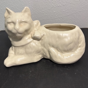 May include: A white ceramic cat planter with a bow around its neck. The cat is lying down with its head turned to the side and its tail curled around its body. The planter has a hole in the top for planting.