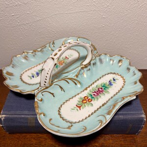 May include: A vintage, two-sectioned serving dish with a handle. The dish is made of white porcelain with a light blue glaze and gold accents. It features hand-painted floral designs in shades of pink, red, yellow, and blue.