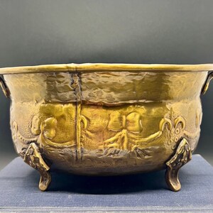 May include: A round brass bowl with a hammered texture, featuring two side handles and three decorative feet. The exterior surface is embossed with a repeating pattern of bows and swags, showcasing a metallic gold finish with dark patina.