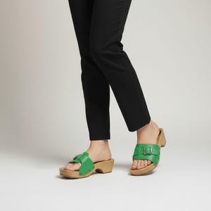 May include: A pair of bright green leather slide sandals featuring a wide strap with a metallic buckle and a light-colored wooden sole. The sandals are worn with slim-fitting black trousers that reach just above the ankle, showcasing the footwear.