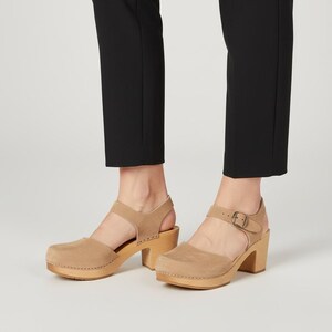 Könnte beinhalten: Beige Wildleder-Clogs mit Holzsohle und hohem Absatz. Die Clogs haben eine geschlossene Spitze, einen Knöchelriemen mit Schnalle und einen Riemen über dem Fuß. Die Schuhe werden mit einer schwarzen Hose getragen.