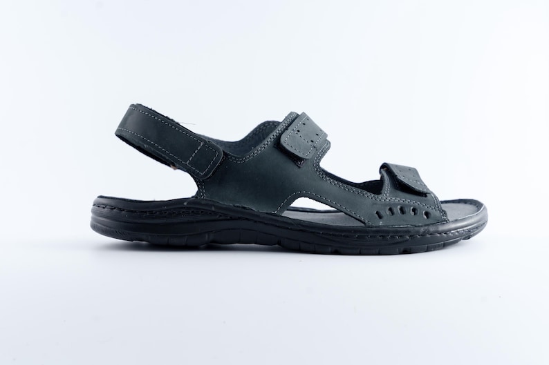 May include: A single dark gray sandal with adjustable straps. The sandal features a textured sole and multiple cut-out details. The straps are secured with hook-and-loop closures. The sandal is designed for comfort and support.