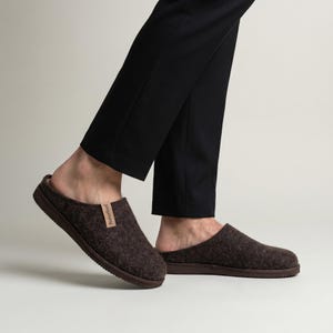 May include: A pair of dark brown felted slippers with a low-back design and a thick, textured brown rubber sole. A small, light tan fabric tag with the brand name Naturfeet is stitched onto the side of the upper part of the footwear.
