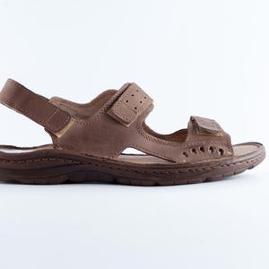 May include: Brown leather sandal with adjustable straps. The sandal features a textured sole and multiple cut-out details. The straps have a hook and loop closure for a secure fit. The sandal is designed for comfort and casual wear.