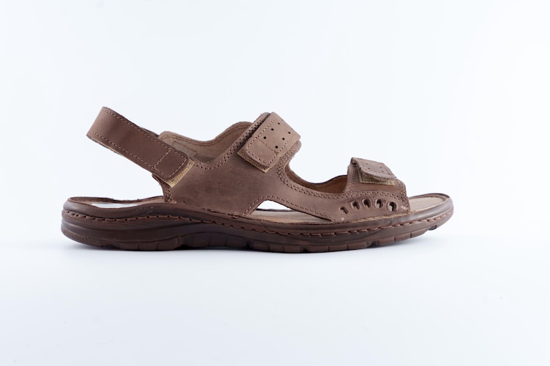 May include: Brown leather sandal with adjustable straps. The sandal features a textured sole and multiple cut-out details. The straps have a hook and loop closure for a secure fit. The sandal is designed for comfort and casual wear.