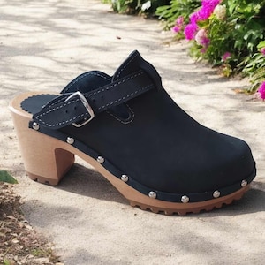 Könnte beinhalten: Schwarzer Clog mit Holzabsatz und Sohle. Der Schuh hat einen Riemen mit silberner Schnalle und ist mit silbernen Nieten um die Sohle verziert. Der Absatz misst etwa 7.6 cm.