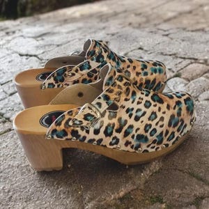May include: A pair of leopard print clogs with blue accents and a buckle. The clogs have a wooden heel and are made of leather.