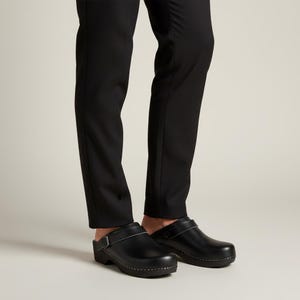 Puede incluir: Un par de zuecos de cuero negro con diseño de punta cerrada, frente redondeado y una correa ajustable en la parte superior asegurada con una hebilla de metal. Los zuecos cuentan con una suela gruesa de color negro sólido y se usan con pantalones negros.